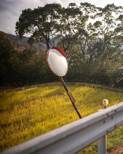 A roadside mirror, facing up towards the sky, reflecting nothing in particular, rice paddy in background.