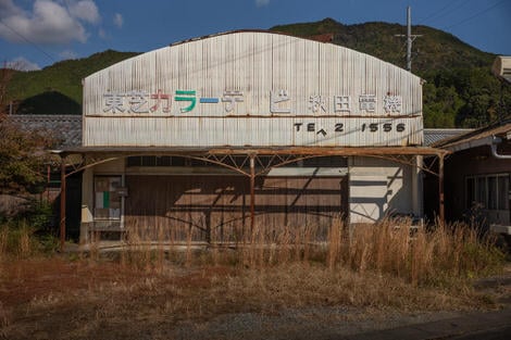 A derelect warehouse or shop or factory for color tvs, signage faded and falling off, weeds in front.