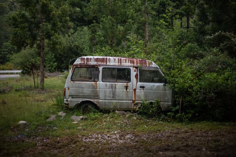 A van, once white, now rust-stained, abandoned in a bush.