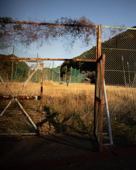A man in a sweatsuit framed by the rusted gate to a field overgrown with weeds.