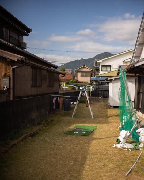 A peek into a residential yard, two ladders converted into a clothes line, shirts and pants drying in the sun, some golf balls siting on a fake green in the foreground.