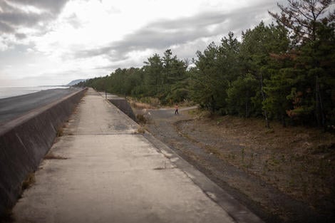Walking atop a seemingly endless seawall, stretching straight towards the horizon, to the right in the far edge of the frame is a man hitting a tennis ball with a racket, mid-swing.