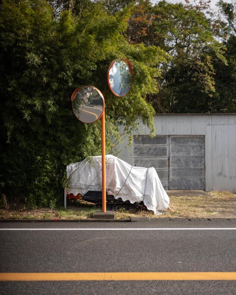 Two roadside mirrors looking in opposite directions, a piece of construction equipment hiding under a white tarp, a corrugated iron shed in the background.